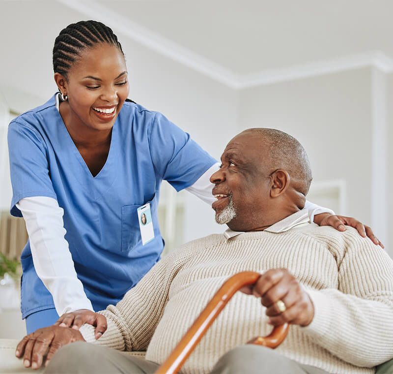 A patient care technician in scrubs helps an older patient sit up in bed, offering support and compassion.